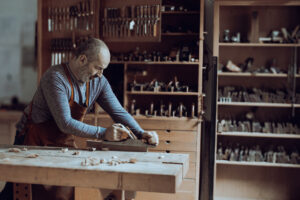 Tom Bensari working at the bench in Bensari Workshop, hand-planing a wooden element using traditional hand tools, surrounded by a wall of woodworking tools.