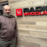 Tom Bensari w siedzibie Radio Wrocław, stojący przy ścianie z logo stacji, przed wejściem do studia nagraniowego