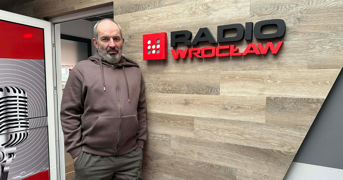 Tom Bensari w siedzibie Radio Wrocław, stojący przy ścianie z logo stacji, przed wejściem do studia nagraniowego